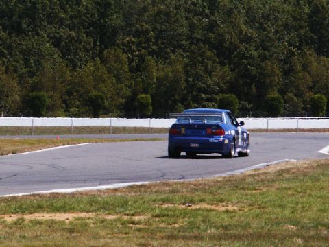 2001 Mercury Grand Marquis - A blue race car navigates a turn on a paved track, with trees lining the background.