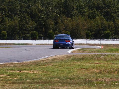 2001 Mercury Grand Marquis - A blue race car navigates a turn on a paved track, with trees and a white barrier in the background.