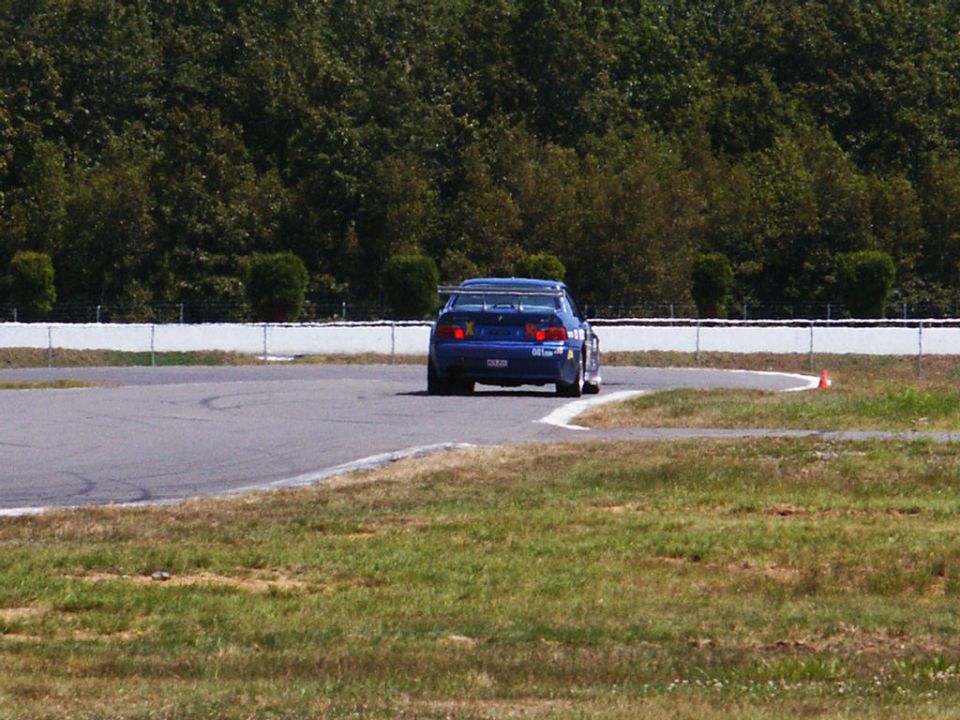 Blue race car with a large rear spoiler on a track, viewed from behind