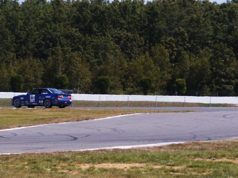 2001 Mercury Grand Marquis - A blue race car navigates a turn on a track, with a dense line of trees bordering the course.