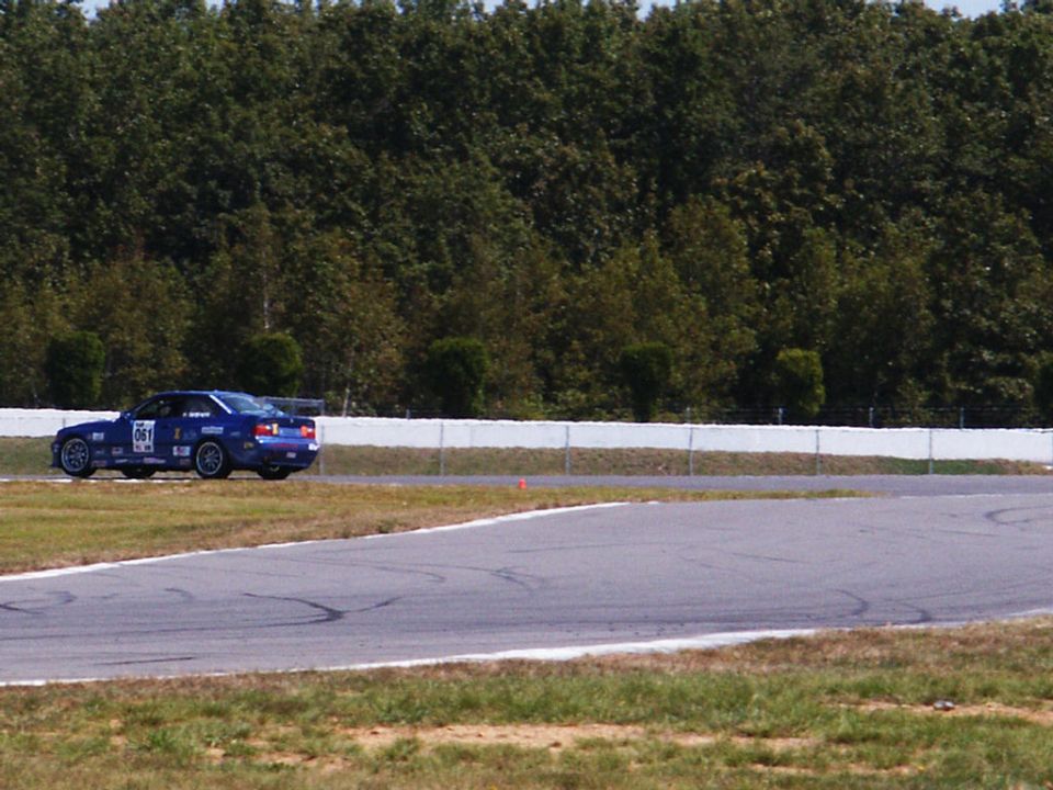 Blue race car with number 061 on door, driving on a paved track with trees in background