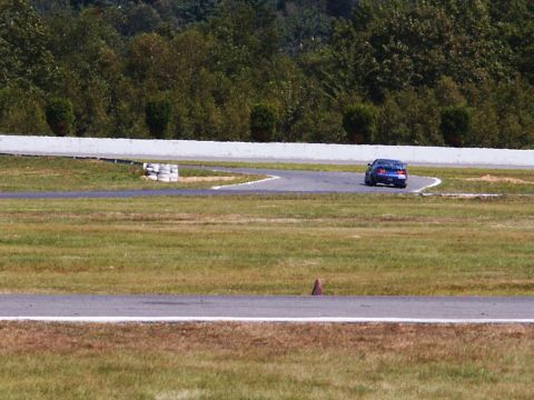 2001 Mercury Grand Marquis - A blue sedan navigates a turn on a race track, with trees and a white barrier in the background.