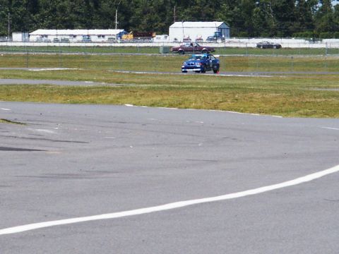2001 Mercury Grand Marquis - A blue race car navigates a turn on a sunny day at the track, with other vehicles visible in the distance.