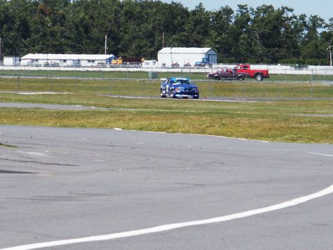 2001 Mercury Grand Marquis - A blue race car drives on a track, with a red pickup truck visible in the distance.