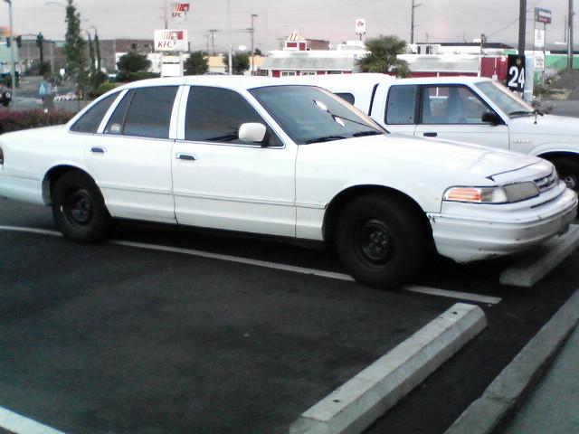 White Ford Crown Victoria sedan parked in a lot, passenger side view, with a white truck and KFC sign behind it.