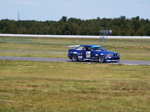 2001 Mercury Grand Marquis - A blue racing car with sponsor decals drives on a paved track with grassy shoulders.