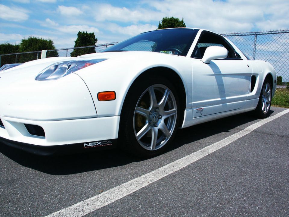 White Acura NSX parked on asphalt with a chain-link fence and trees in the background