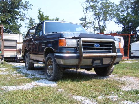 1993 Ford Bronco - A dark blue Ford Bronco with a brush guard is parked on a gravel lot with trees in the background.