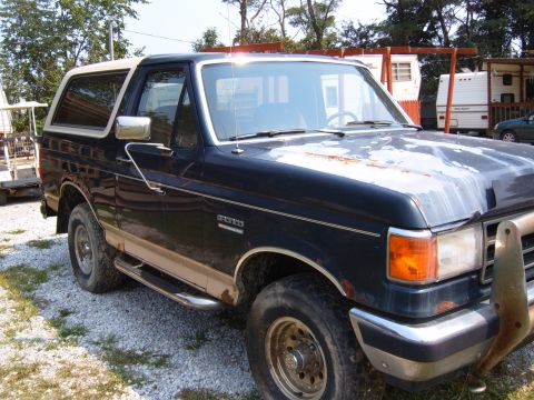 1993 Ford Bronco - A dark blue and tan Ford Bronco with a white roof is parked on a gravel lot, showing signs of wear and tear.