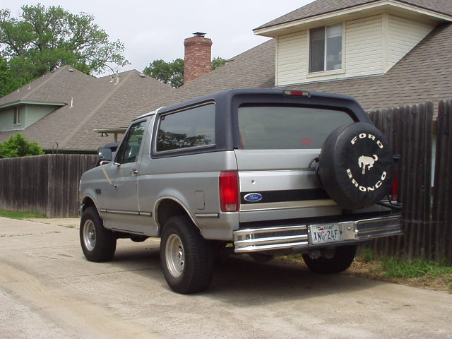 Silver Ford Bronco SUV from rear driver side with Ford Bronco spare tire cover, parked by a wooden fence