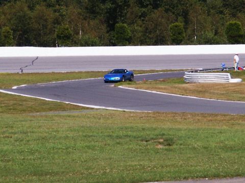 2001 Mercury Grand Marquis - Blue sports car navigates a turn on a race track with trees and a white wall in the background.