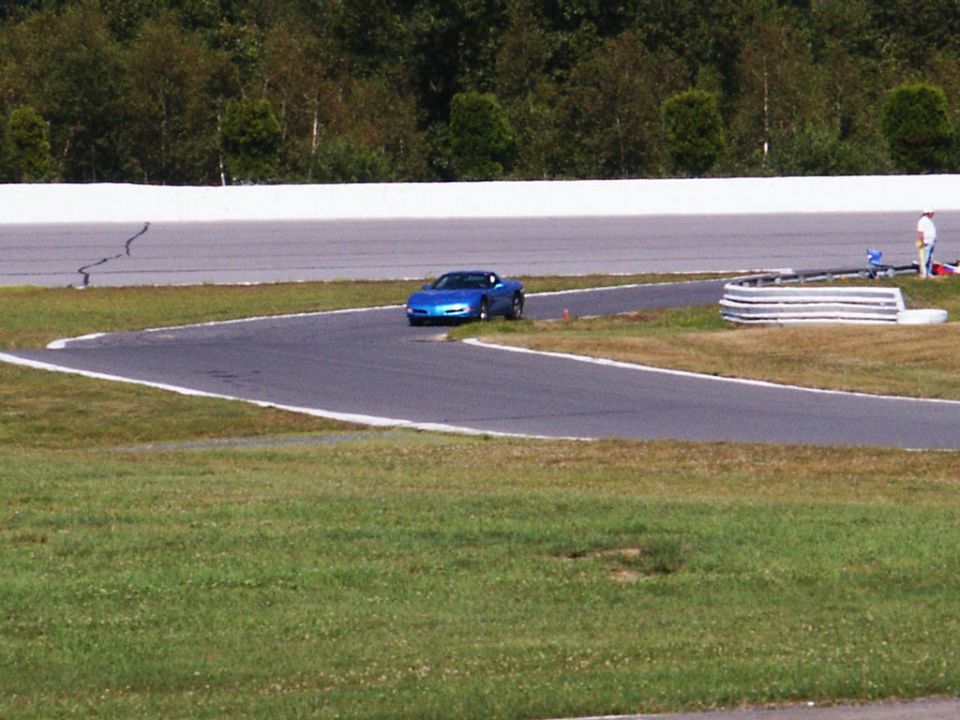 Blue sports car on a race track corner. A person stands near a barrier in the background