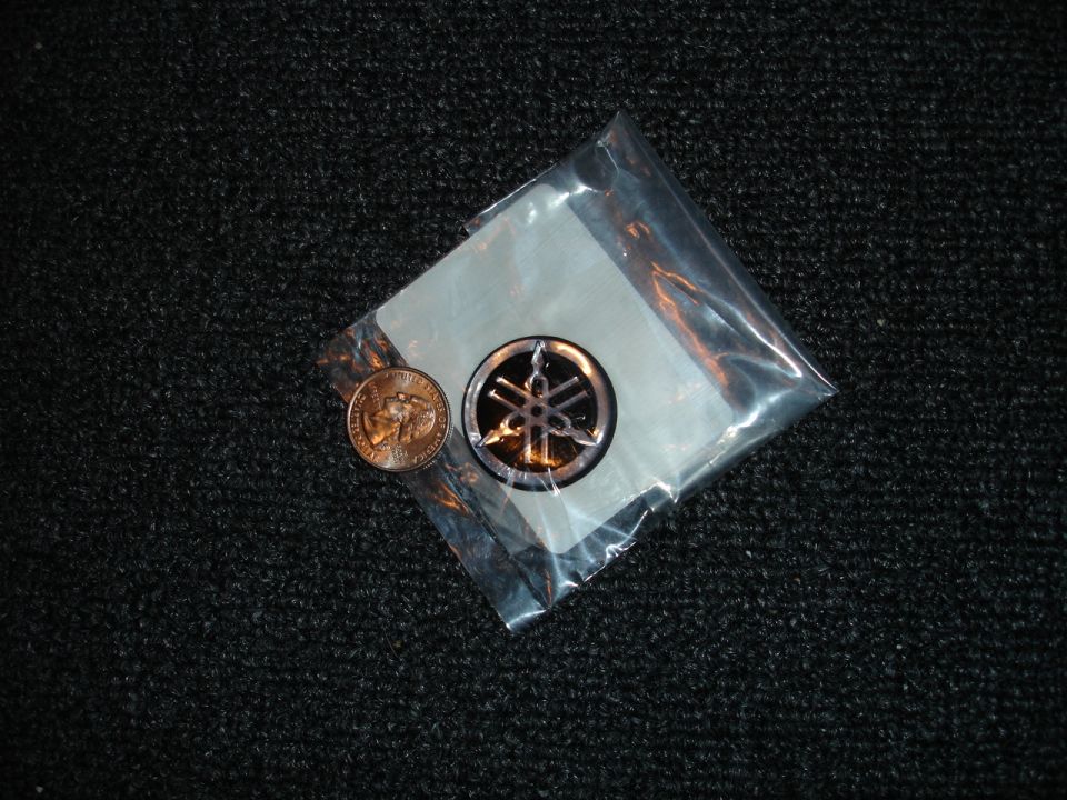 Yamaha emblem in plastic packaging next to a penny on a black textured surface