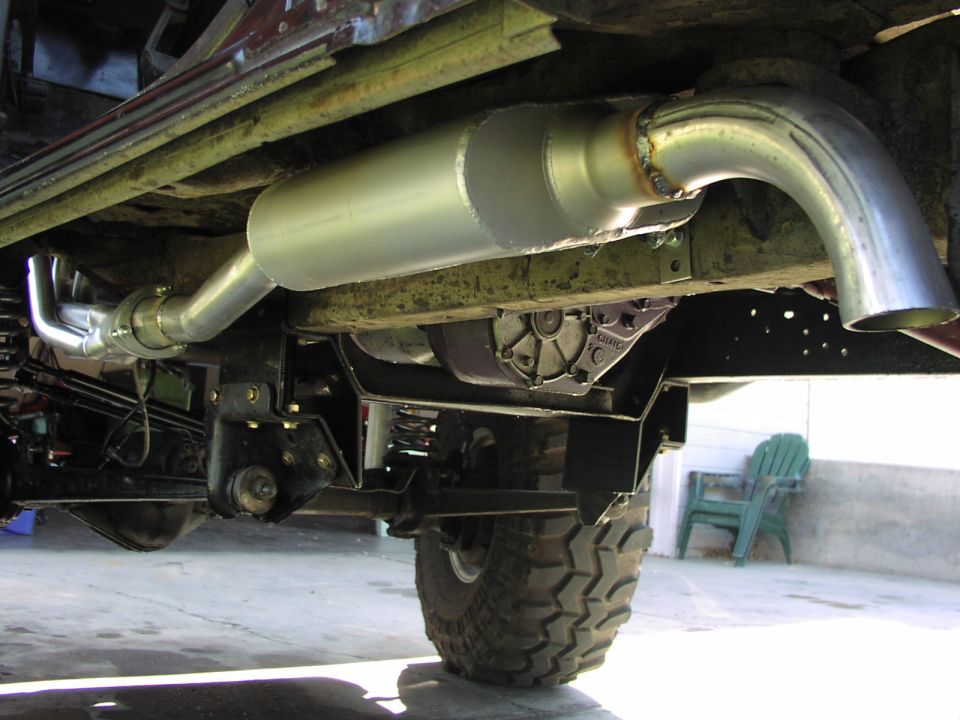 Underside view of a vehicle's exhaust system, including headers, muffler, and tailpipe, with drivetrain components like the transmission visible