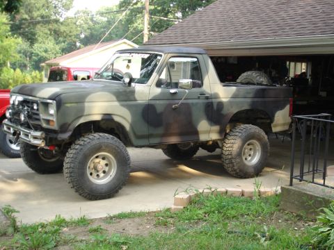 1979 Ford Bronco - Lifted, camouflaged pickup truck with off-road tires sits in a driveway next to a garage.