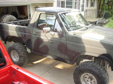 1979 Ford Bronco - A camouflaged SUV is parked in a driveway next to a red truck. The SUV has a lifted suspension and large off-road tires.