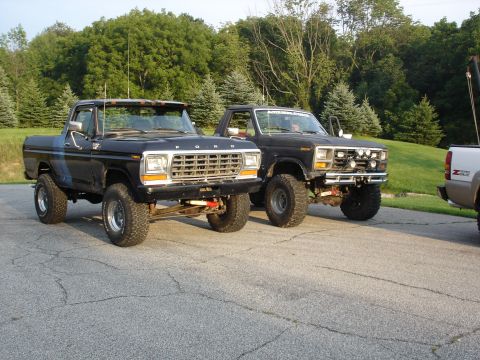 1979 Ford Bronco - Two lifted Ford trucks, including a 1979 Bronco, are parked on a paved surface with trees in the background.