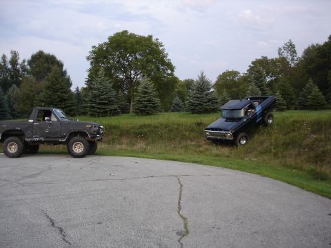 1979 Ford Bronco - Two lifted pickup trucks are parked on a paved surface next to a grassy hill with trees in the background.
