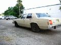 1997 Ford Crown Victoria - A tan sedan is parked in a gravel lot next to a building. Another vehicle is visible in the background.