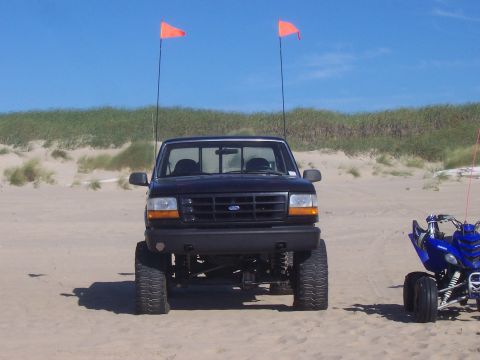 1994 Ford F-150 - A lifted black Ford pickup truck sits on a sandy beach, with two orange flags flying high against a clear blue sky.