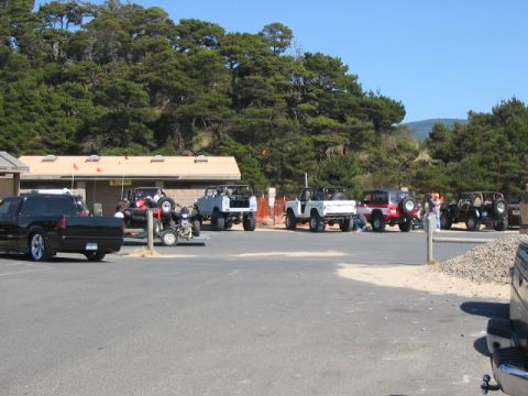 1995 Ford Bronco - A group of off-road vehicles are parked in a lot near a wooded area and a building on a sunny day.
