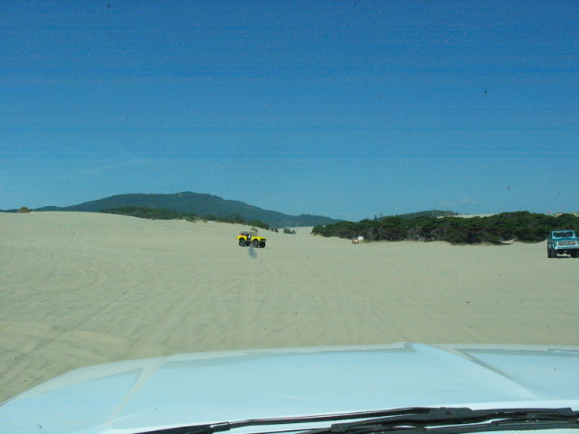 Dashboard POV of sand dunes with distant yellow and blue vehicles under blue sky