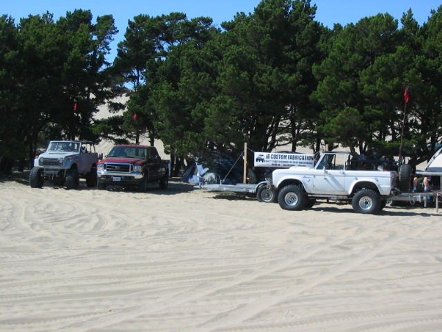 White lifted early Ford Bronco parked on sand with other 4x4s and trees behind.