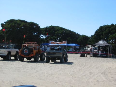 1995 Ford Bronco - Several off-road vehicles are parked on a sandy beach, with tents and trees in the background under a clear blue sky.
