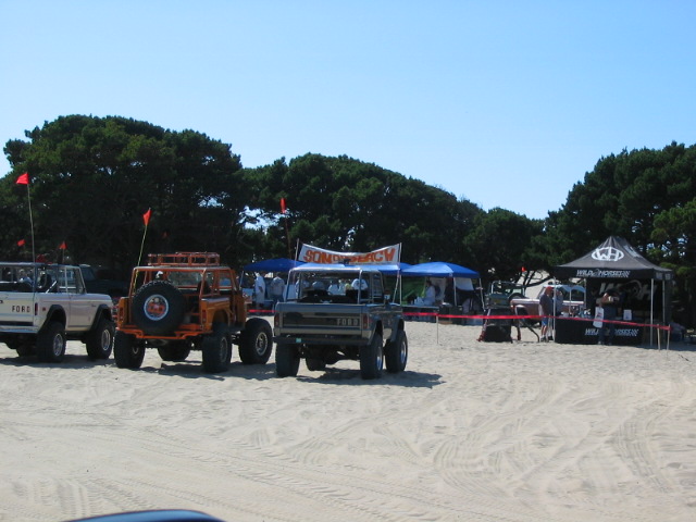 Early Ford Broncos parked on sand at an off-road beach event with tents and flags