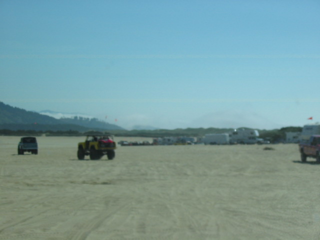 Vehicles including a light SUV and yellow off-road vehicle on a sandy beach with RVs in the distance