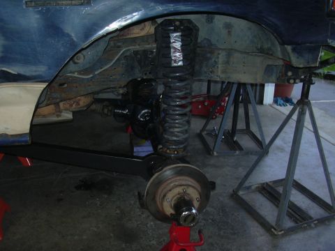 1991 Ford Bronco - Front suspension detail of a blue truck up on jack stands in a garage with the wheel and inner fender removed, showing a tall coilover shoc…