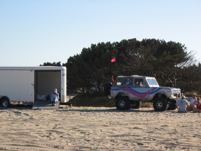 Lifted white Ford Bronco with pink/purple graphics parked on sand beside an open enclosed trailer