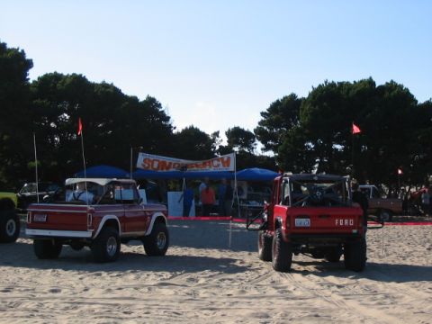 1995 Ford Bronco - Two red Ford Broncos parked on a sandy beach with a banner in the background.