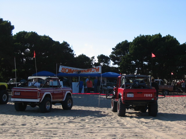 Two red Ford Broncos parked on beach sand at an event with tents and flags