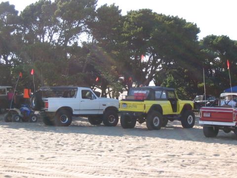 1995 Ford Bronco - Several off-road vehicles are parked on a sandy beach near trees.