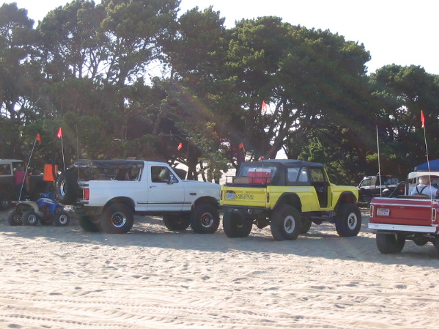 Lifted 4x4 trucks and a yellow Bronco parked on a beach with tall safety flags