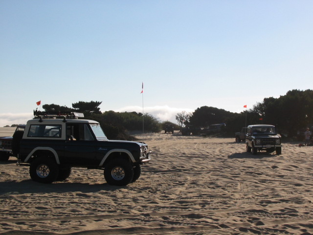 Dark early Ford Bronco with white fenders and big tires parked on sand dunes