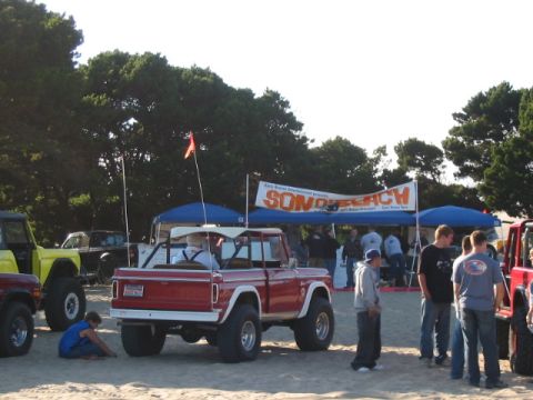 1995 Ford Bronco - A red convertible SUV with a white top is parked on the sand near an event tent with a "SONO BEACH" banner.