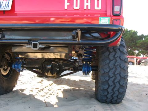 1995 Ford Bronco - Close-up of the rear axle and suspension of a red Ford truck, showing off-road modifications and large tires on a sandy surface.