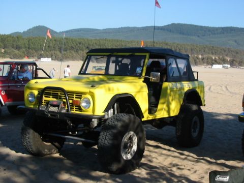 1995 Ford Bronco - A lifted yellow Ford Bronco sits on a sandy beach with dunes and trees in the background.