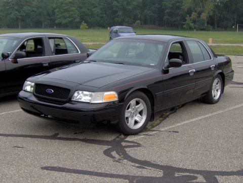 2002 Ford Crown Victoria - Two black Ford Crown Victorias are parked in a lot. The front vehicle is angled towards the camera.