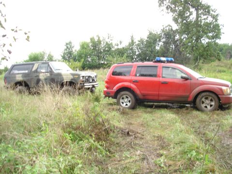 1979 Ford Bronco - A camouflaged Ford Bronco appears stuck in the mud, with a red SUV equipped with a light bar positioned behind it.