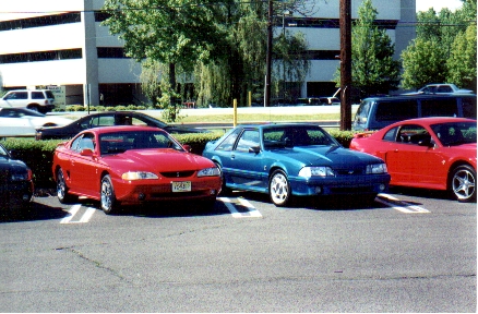 1989 Ford Mustang - Three cars are parked in a lot, including a red Mustang and a blue Mustang Fox Body.