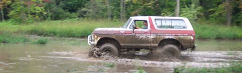 1979 Ford Bronco - A red and white Ford Bronco navigates a muddy path, kicking up water and dirt.