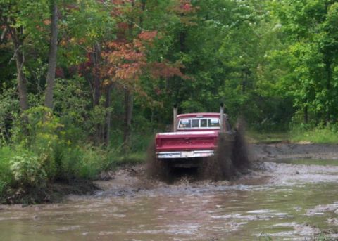 1979 Ford Bronco - Red pickup truck navigates a muddy trail, splashing water and dirt.
