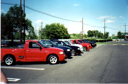 1989 Ford Mustang - A line of pickup trucks are parked in a lot under a bright blue sky.