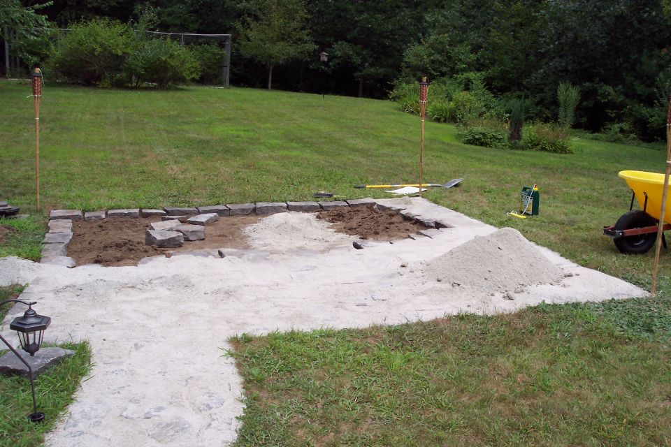 Patio construction site with sand, gravel, stones, and a yellow wheelbarrow