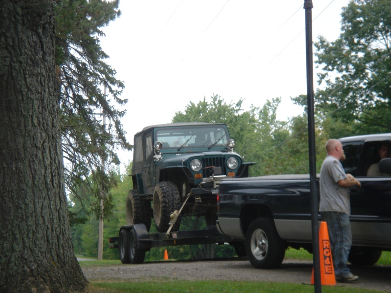 Green Jeep Wrangler on a trailer, towed by a dark pickup truck with a man standing by the driver's door