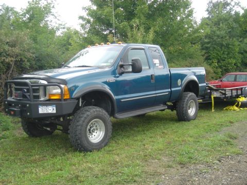 1979 Ford F-250 - A blue pickup truck tows a trailer carrying a red car, equipped with a heavy-duty aftermarket bumper.