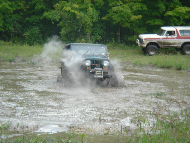 Green Jeep Wrangler splashing mud in a muddy field with a white and red Ford Bronco in the background